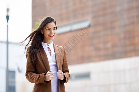 轻的女人,留着漂亮的头发,站城市背景里穿正式服装的女商人,留着波浪式发型穿着棕色夹克裤子的轻女孩图片