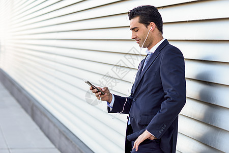 轻的商人穿着蓝色西装领带,城市背景下用智能手机街上穿着正式衣服耳机的男人图片