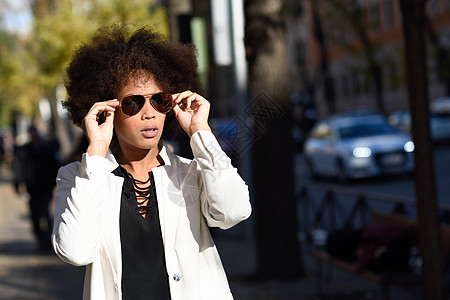 轻的黑人妇女,留着AFRO发型,站城市背景下,戴着飞行员太阳镜穿着白色夹克黑色连衣裙的混合女孩砖墙附近摆姿势图片