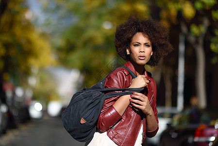 轻的黑人女,留着AFRO发型,站条城市街道上,背着个包混合女人穿红色皮夹克白色连衣裙与城市背景背景图片
