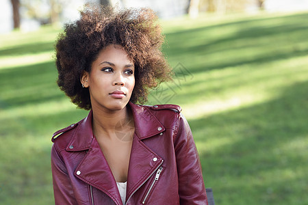 轻的黑人女,留着AFRO发型,坐城市公园的长凳上混合女人穿红色皮夹克白色连衣裙与城市背景图片