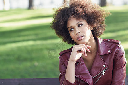 轻的黑人女,留着AFRO发型,坐城市公园的长凳上混合女人穿红色皮夹克白色连衣裙与城市背景背景图片