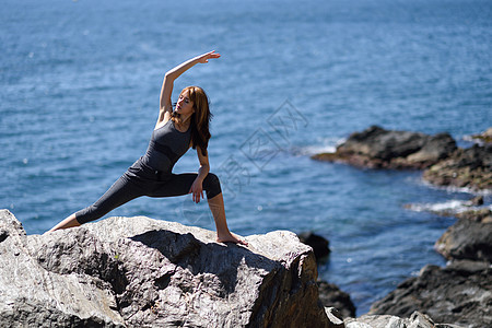 轻的女人海滩上瑜伽女穿着运动服,岩石上穿运动服,背景海图片