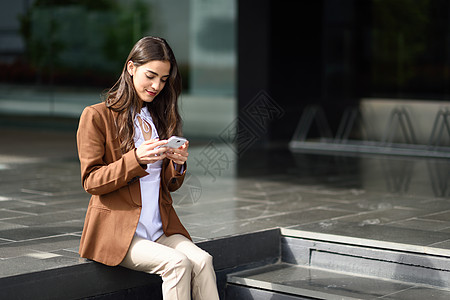 轻的女商人办公楼旁边用智能手机发短信穿着正式服装的美女用智能手机轻女孩,棕色夹克城市背景图片