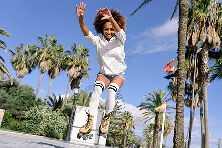 穿着溜冰鞋的轻黑人女人海滩附近跳跃阳光明媚的长廊上留着AFRO发型的女孩背景图片