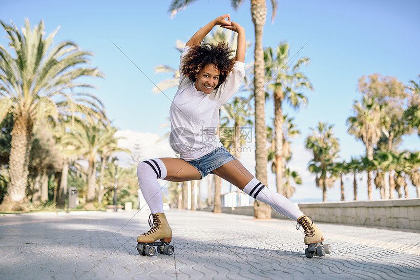 微笑的黑人妇女溜冰鞋骑户外海滩长廊与棕榈树阳光明媚的日子里,带着AFRO发型的微笑女孩穿着溜冰鞋的黑人女人海滩长廊图片