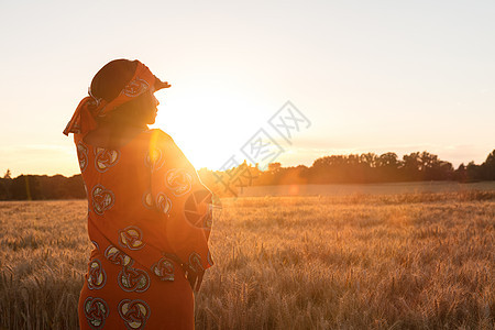 非洲妇女穿着传统的衣服,站着,看着,手着眼睛,大麦小麦作物的田野日落日出图片