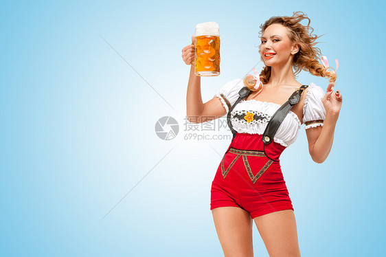 轻的调情感的瑞士女人穿着红色跳线短裤吊带的传统Dirndl,着蓝色背景的啤酒杯图片