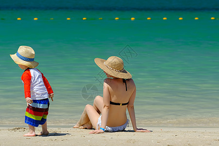 两岁的蹒跚学步的男孩戴着太阳帽,妈妈海滩上图片