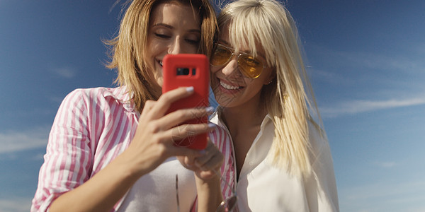 秋天的天,两个女孩朋友空荡荡的海滩上用智能手机拍照图片
