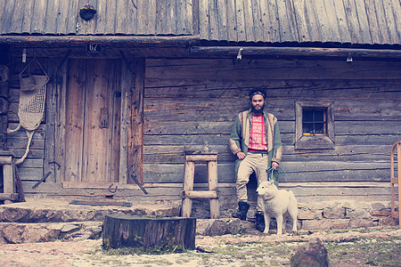 英俊的轻时髦男子白色哈士奇狗站,古老的复古木屋前木屋前带着狗的嬉皮士图片