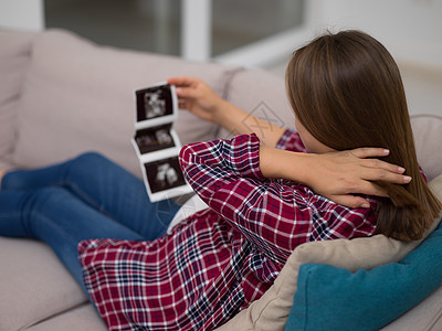 孕妇看着婴儿的超声波轻孕妇看着婴儿的超声波照片,同时家里的沙发上放松图片