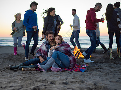 轻的夫妇日落时朋友坐海滩上的篝火旁喝啤酒夫妇海滩上日落时朋友享受图片