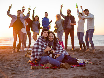 轻的夫妇日落时朋友坐海滩上的篝火旁喝啤酒夫妇海滩上日落时朋友享受图片