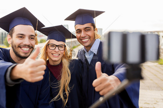 教育毕业技术人的群快乐的国际学生穿着砂浆板学士服,户外用智能手机自拍,竖大拇指学生毕业生用智能手机自拍图片