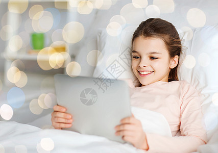 人,孩子技术快乐的微笑女孩躺床上的平板电脑电脑家里的假日灯光背景快乐的女孩床上用平板电脑超过灯光图片