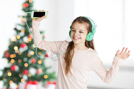 假期,儿童技术快乐的微笑女孩戴着耳机,带着智能手机,诞树背景上听音乐跳舞诞节智能手机耳机的女孩图片