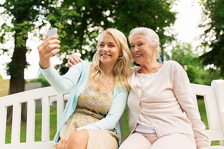家庭,技术人的快乐微笑的轻女儿资深母亲与智能手机坐公园长凳上自拍女儿老母亲公园自拍图片