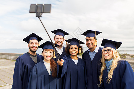 教育毕业技术人的群快乐的国际学生穿着迫击炮板学士服,用智能手机自拍杆户外拍照群快乐的学生毕业生自拍图片