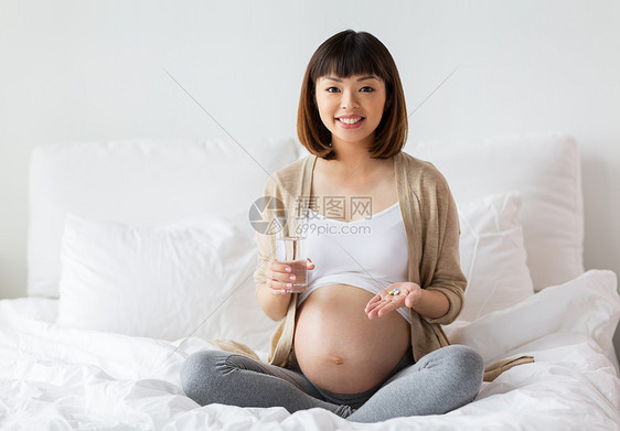 怀孕,人保健快乐的亚洲孕妇坐家里的床上,着药丸杯水快乐的孕妇床上吃药家图片