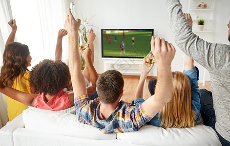友谊休闲人的快乐的朋友与含酒精的啤酒坐沙发上,家里看足球足球比赛啤酒的朋友家里看电视上的足球图片