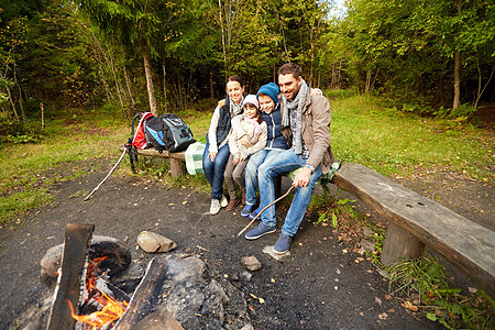 野营,旅游,徒步旅行人们的快乐的家庭坐长凳上的营地火灾树林里幸福的家庭坐篝火旁的长凳上幸福的家庭坐篝火旁图片