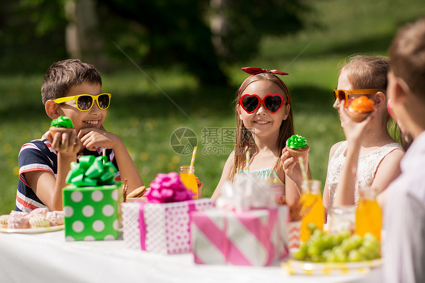 假期,童庆祝快乐的孩子戴着太阳镜坐桌子上的生日聚会夏季花园吃纸杯蛋糕孩子们夏天的生日聚会上吃纸杯蛋糕孩图片