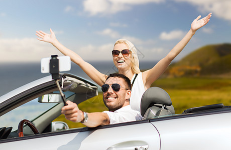 公路旅行,技术旅行快乐的夫妇驾驶敞篷汽车,并智能手机自拍杆拍照幸福的夫妇汽车上用智能手机自拍图片