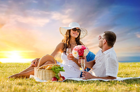 爱情,约会,人庆祝的微笑的夫妇与鲜花喝香槟草地上野餐日落天空背景夫妇带着鲜花野餐时喝香槟图片