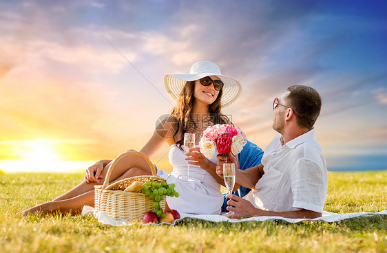 爱情,约会,人庆祝的微笑的夫妇与鲜花喝香槟草地上野餐日落天空背景夫妇带着鲜花野餐时喝香槟图片