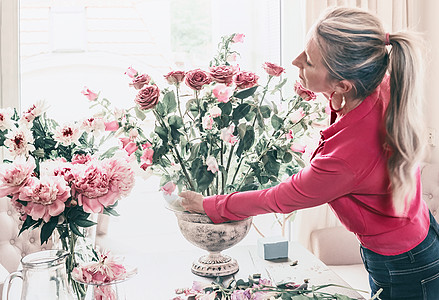 花店妇女穿着红色衬衫,制作美丽的大型节日活动,古典花与玫瑰其他花骨灰瓶桌子上的窗口,生活方式图片