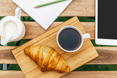 早上早餐绿色花园与法国牛角包,咖啡杯,橙汁,平板电脑笔记簿木桌上图片