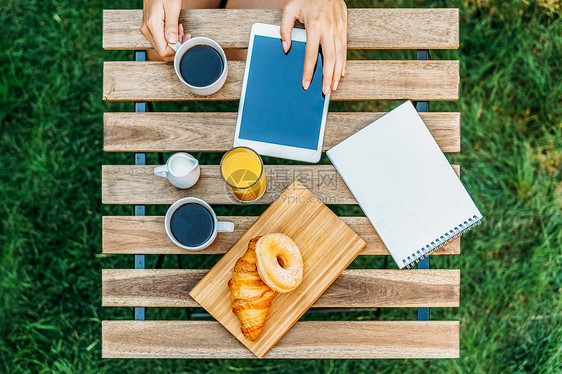 轻而魅力的女人绿色花园里吃早餐,法国牛角包甜甜圈咖啡杯橙汁平板电脑木制桌子上的笔记簿图片