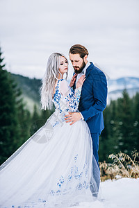 新郎穿着蓝色的西装,新娘穿着白色的衣服,绣着蓝色的图案,穿着喀尔巴阡山脉绿色松林的背景上图片