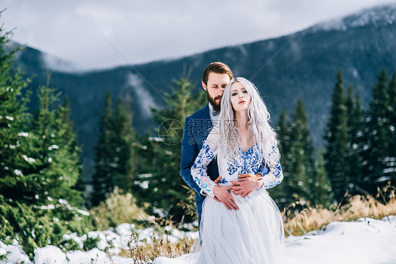 新郎穿着蓝色的西装,新娘穿着白色的衣服,绣着蓝色的图案,穿着喀尔巴阡山脉绿色松林的背景上图片