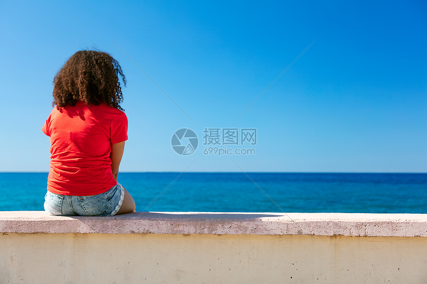 混合种族非裔美国女孩十几岁的女轻女子穿着红色T恤牛仔短裤坐墙上,向外望着蓝色的大海图片