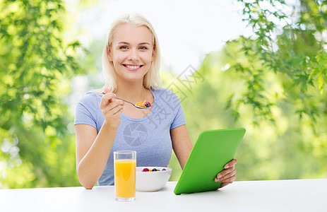 健康的饮食,食物技术微笑的轻妇女与平板电脑,穆斯利果汁早餐绿色自然背景微笑的女人用平板电脑吃早餐图片