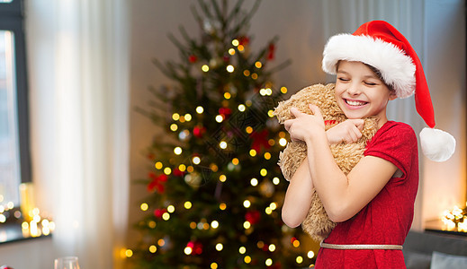 童假期的微笑的女孩诞老人帽子与泰迪熊房间与诞树背景诞节戴着泰迪熊的诞老人帽子的女孩图片