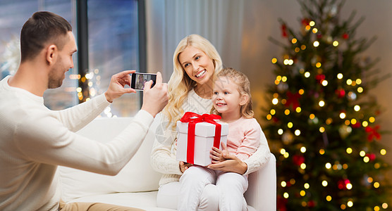 假期,技术人的快乐的父亲家里用智能手机拍摄女儿AMD母亲的照片,背景诞树灯诞节给家人拍照的男人图片