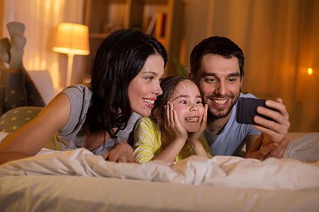 人们家庭的快乐的母亲,父亲小女儿,晚上家里睡觉时带着智能手机快乐的家庭,晚上床上智能手机图片