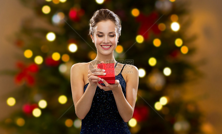 节日,奢侈人的微笑的女人穿着衣服,着红色的礼品盒诞树的灯光背景诞树灯上红色礼品盒的女人图片