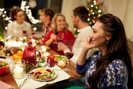 假期,沟通庆祝快乐的轻妇女打电话给智能手机,并与朋友家里吃诞晚餐诞节晚餐时打电话给智能手机的女人图片