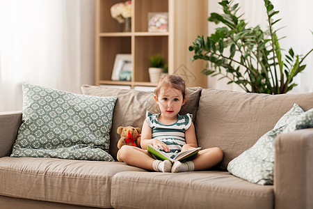 童人们的快乐的小女孩坐沙发上,带着书玩具泰迪熊家快乐的女孩坐沙发上,家里书图片