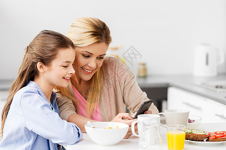 家庭,技术人的快乐的母亲女儿与智能手机家里的厨房吃早餐智能手机的家庭家吃早餐图片