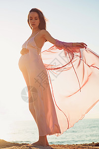 穿着粉红色连衣裙的美丽孕妇的户外大气生活方式肖像美好的早晨海滩上的日出美丽时尚图片