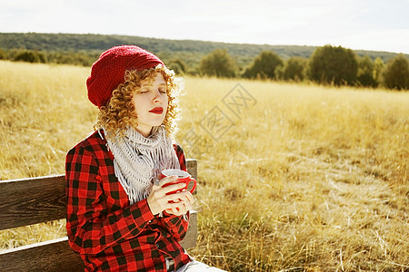 位穿着红色格子衬衫戴着羊毛帽围巾的轻女子的正肖像,她正日光浴,坐黄色田野的木凳上,背光来自秋天的阳光图片