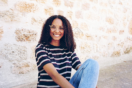 幅幸福微笑的轻黑人妇女的肖像,她戴着眼镜牛仔裤件条纹t恤,坐地上,卷发图片