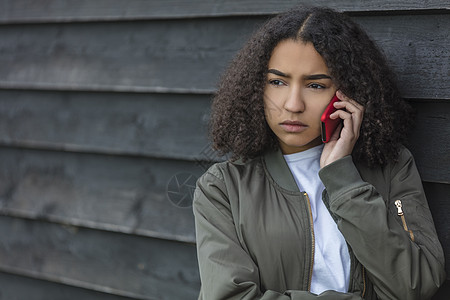 户外肖像美丽,悲伤,侵略,混合种族,非裔美国女孩,十几岁的女青,红色手机上交谈,穿着绿色轰炸机夹克图片