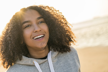 户外肖像美丽的快乐混合种族非裔美国女孩十几岁的女轻女子海滩上微笑着用完美的牙齿金色的日落黄昏图片