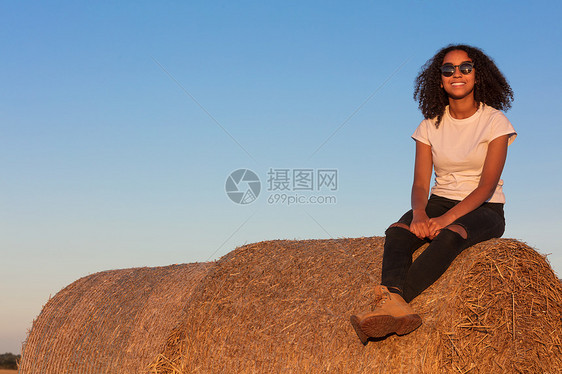 户外肖像美丽的快乐混合种族非裔美国女孩十几岁的女轻女子笑着与完美的牙齿太阳镜坐干草甘蓝金色的傍晚阳光图片